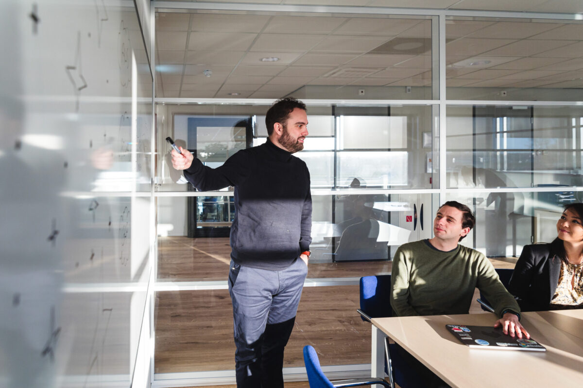 A person in front of a whiteboard pointing to it and two people in the same meeting room listing to him.