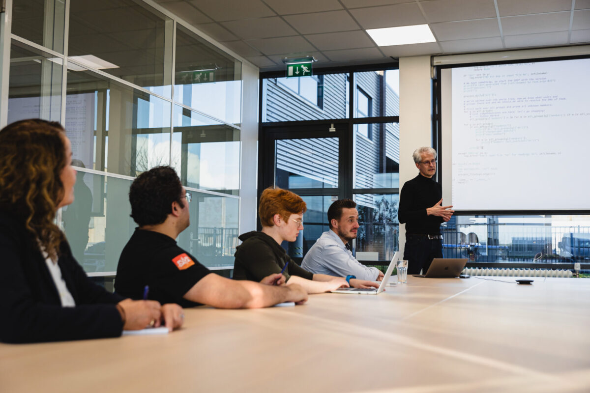 Five people in a meeting room. One stands in front of a screen with code and is explaining.