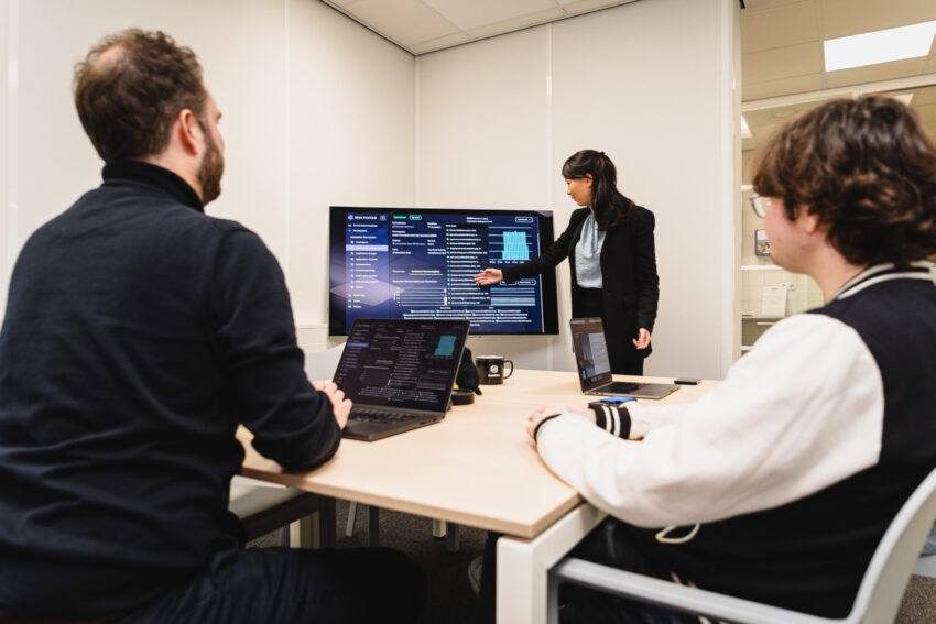 Three people in a meeting room. One is standing in front of a screen presenting.