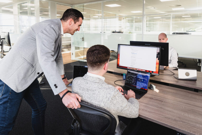 A person working on a computer with someone standing next to him. In the background there is another person behind a screen.